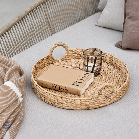 Flettet rundt bakke på lysegrå sofa med en bog med titlen FOSS og en glaslysestage med lys, ved siden af et beige stribet tæppe og puder.