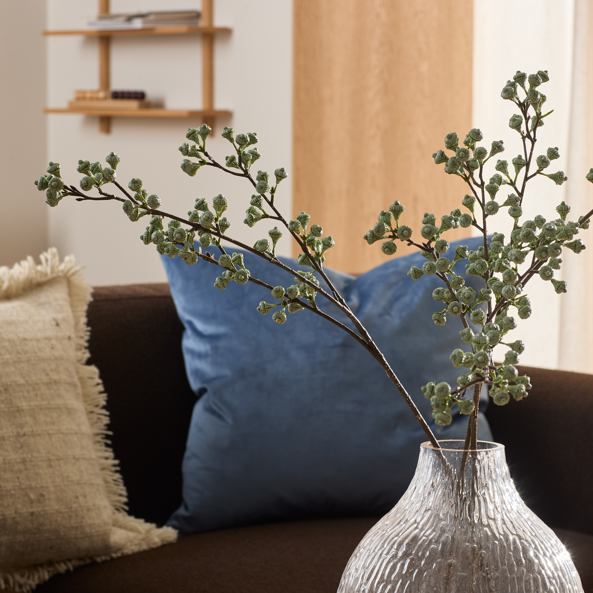 Glasvase med to grønne kviste i fokus, brun sofa med blå og beige pude i baggrunden i en lys stue.