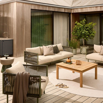 Lys terrasse med beige loungemøbler i flettet look, træbord på rundt tæppe og krukplanter foran store glasdøre.