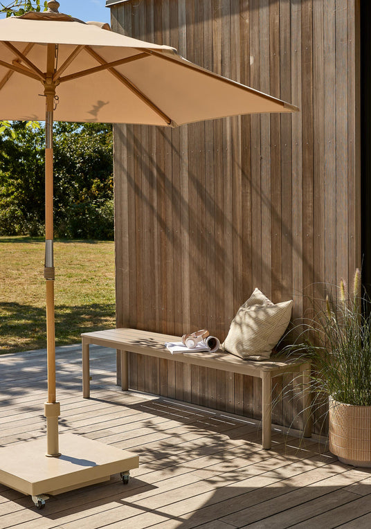 Solrig terrasse med beige parasol, træbeklædt væg og bænk med pude og rullet magasin; krukkeplante ved siden af.