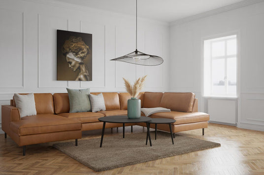 Lyst stue med brun lædersofa, sorte sofaborde, pampasgræs i grøn vase, moderne pendellampe og vægpaneler ved vindue i dagslys.
