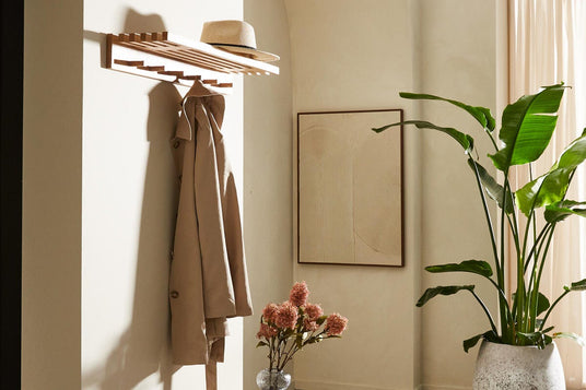 Lys entré med træbænk, vægmonteret hylde med beige frakke og stråhat, stor grøn plante og glasvase med blomster.