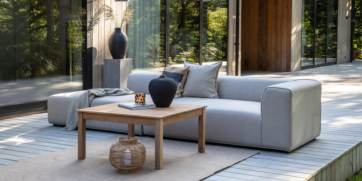 Moderne terrasse med lysegrå sofa, træbord og vaser foran et træhus med store glaspartier i dagslys.
