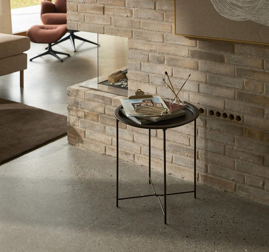 Lille sort sidebord med magasiner og pensler i et glas foran en lys murstenspejs i en moderne stue.