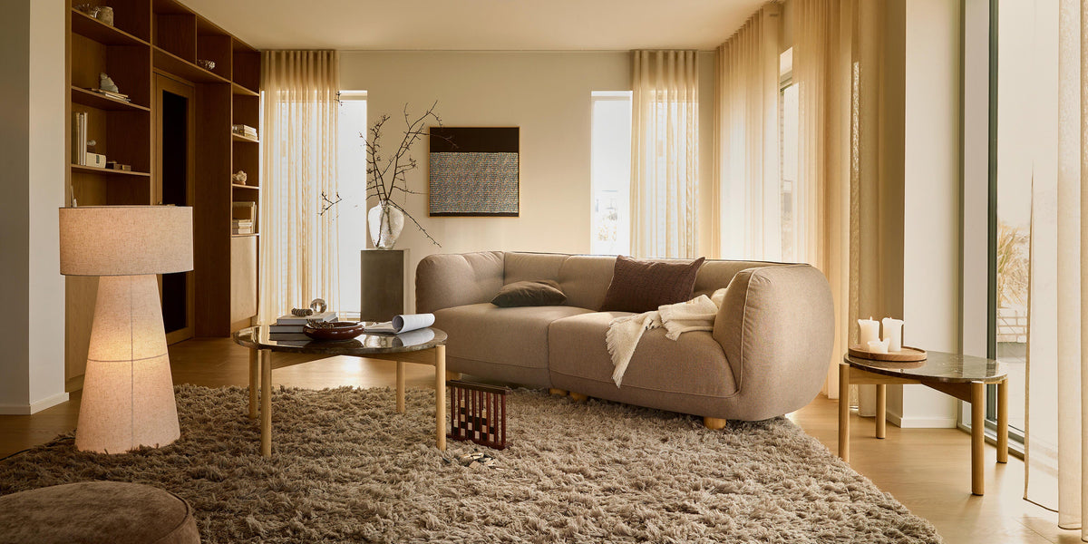 Lys stue i beige nuancer med buet sofa, blødt ryatæppe, gulvlampe, to borde med stearinlys og lette gardiner ved store vinduer.