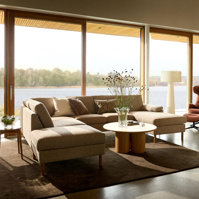 Lys stue med beige modulsofa, rundt sofabord i træ og store panoramavinduer med udsigt over en sø.