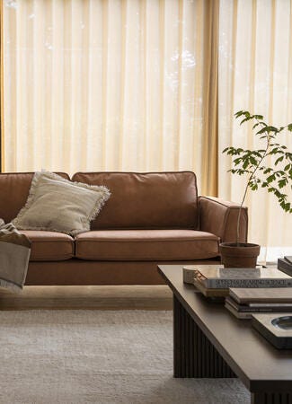 Lyst stue med brun lædersofa, frynset pude og plaid, beige gardiner, en potteplante og et lyst tæppe på trægulv i dagslys.