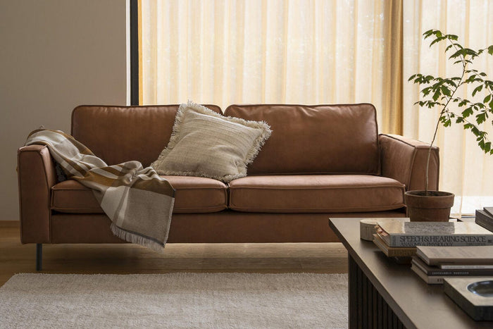 Lyst stue med brun lædersofa, frynset pude og plaid, beige gardiner, en potteplante og et lyst tæppe på trægulv i dagslys.