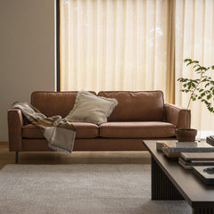 Lyst stue med brun lædersofa, frynset pude og plaid, beige gardiner, en potteplante og et lyst tæppe på trægulv i dagslys.