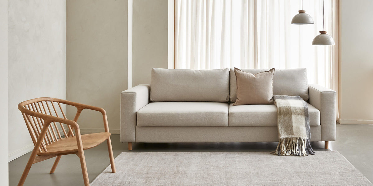 Lyst, minimalistisk stue med beige sofa med ternet plaid og pude, træstol, to pendellamper, lyse gardiner og stort blødt tæppe.