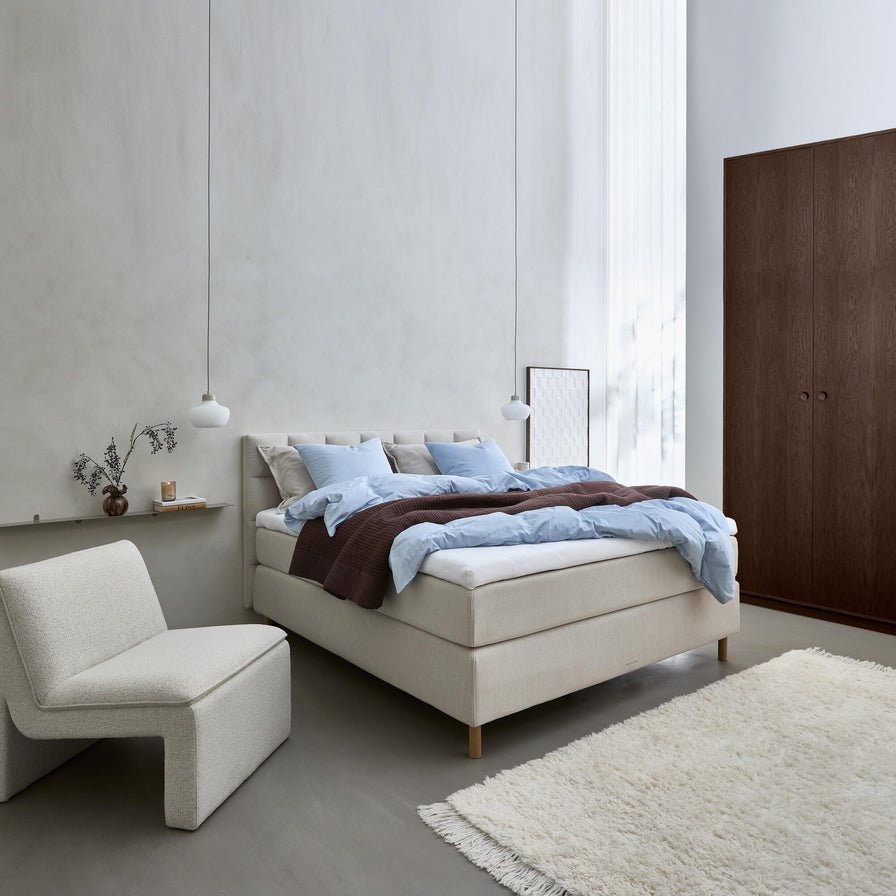 Lyst skandinavisk soveværelse med beige polstret seng, lyseblåt sengetøj og brun plaid, to pendler, lav lounge-stol, vase med grene på hylde, lette gardiner, hvid langhåret tæppe og mørkt klædeskab.