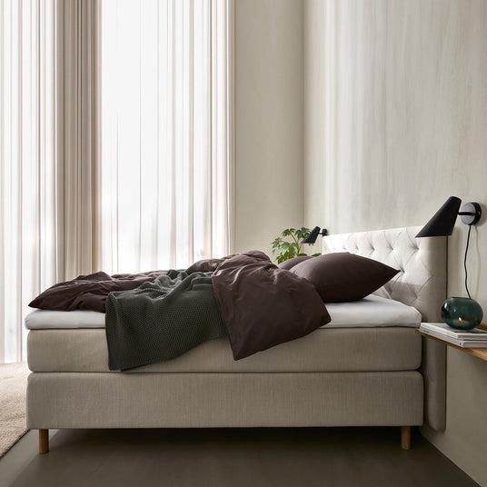 Lyst soveværelse med beige kontinentalseng og polstret sengegavl, mørkebrunt sengetøj og grøn plaid, sorte væglamper, lette gardiner samt lille hylde med glasvase og bøger.