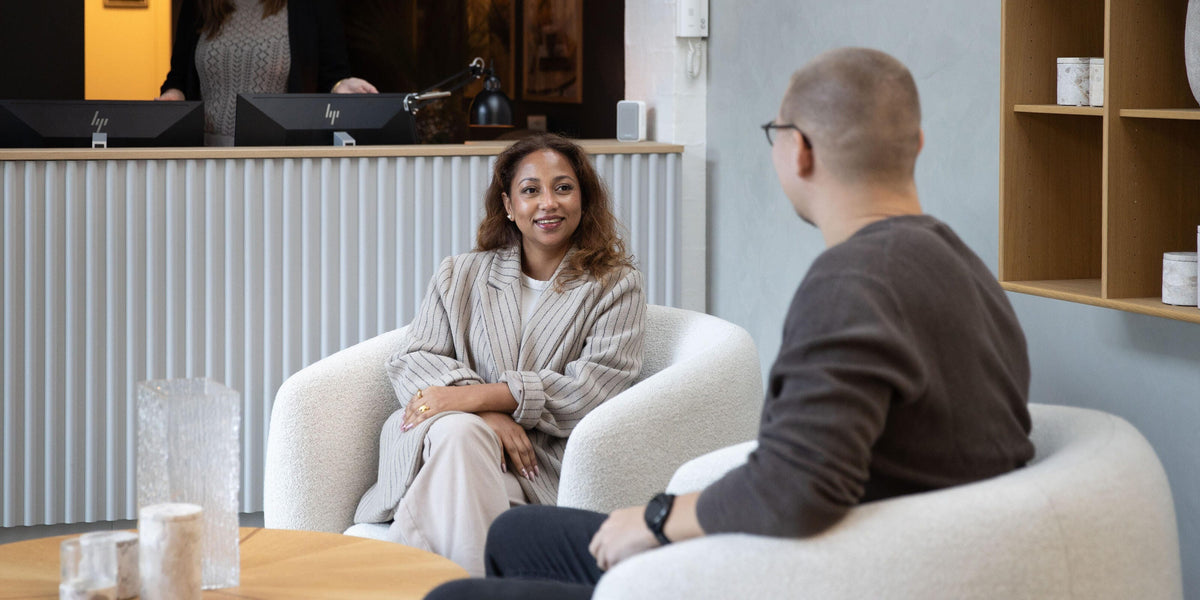 Moderne showroom-reception med to personer i hvide lænestole og rundt træbord med glasvaser; på væggen står “Welcome home – We believe everyone deserves a place they feel at home”.