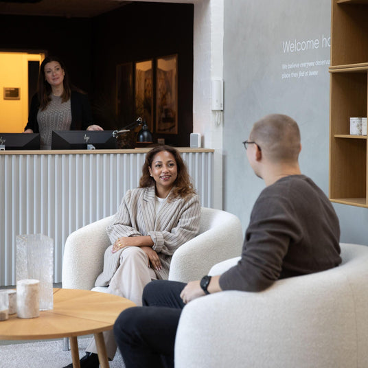Moderne showroom-reception med to personer i hvide lænestole og rundt træbord med glasvaser; på væggen står “Welcome home – We believe everyone deserves a place they feel at home”.