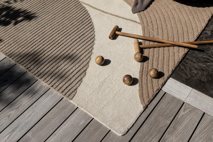 Beige og taupe abstrakt tæppe på en træterrasse med buet sofa, plaid samt trækøller og kugler i sollys.