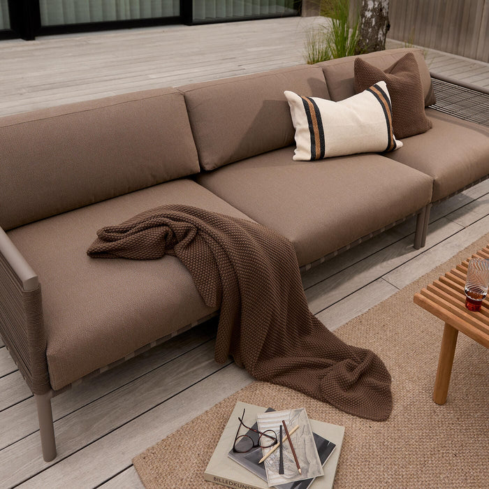 Brun udendørs sofa på terrasse med puder, strikket plaid og et træbord med glas og magasiner.