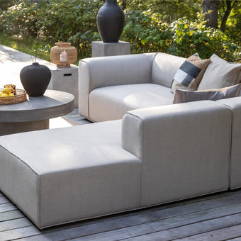 Lysgrå modulsofa med chaiselong på træterrasse, rundt betonbord med sort vase og bakke med citroner; puder og fletlanterne, omgivet af grønne træer i dagslys.