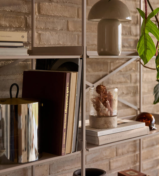 Nærbillede af reolsystem med bøger, hvor en har titlen ”the soft furnishings source book” og en anden ”SOFT MINIMAL”, lille bordlampe, glasvase med tørrede planter, grøn plante og metalbeholder mod murstensvæg.