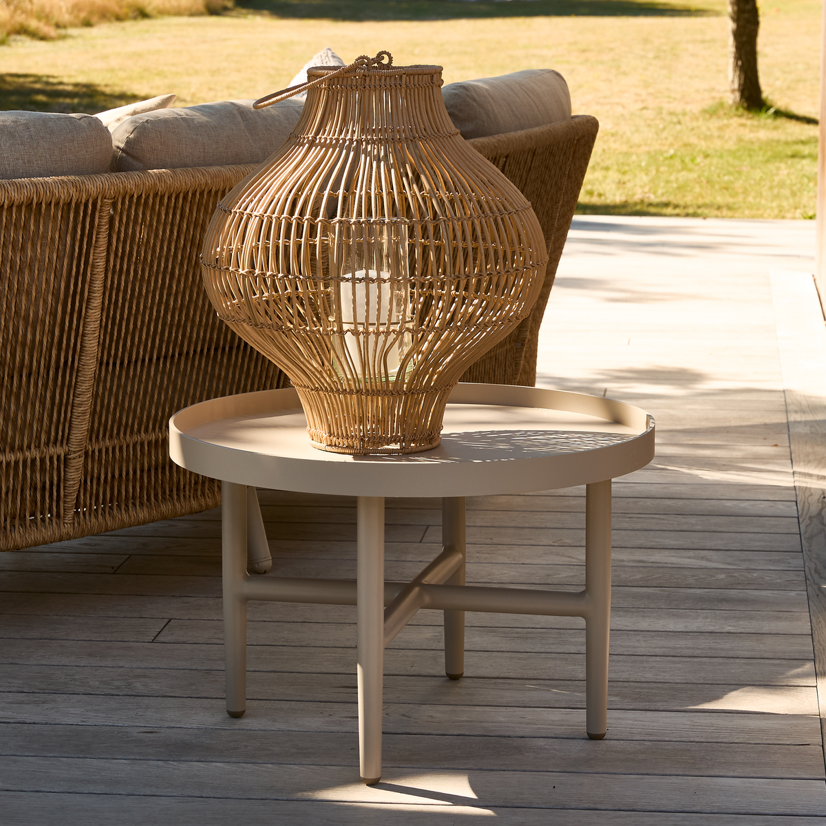 Rundt beige sidebord på træterrasse med stor rattanlanterne ovenpå, udendørs i sollys ved siden af en kurvesofa.
