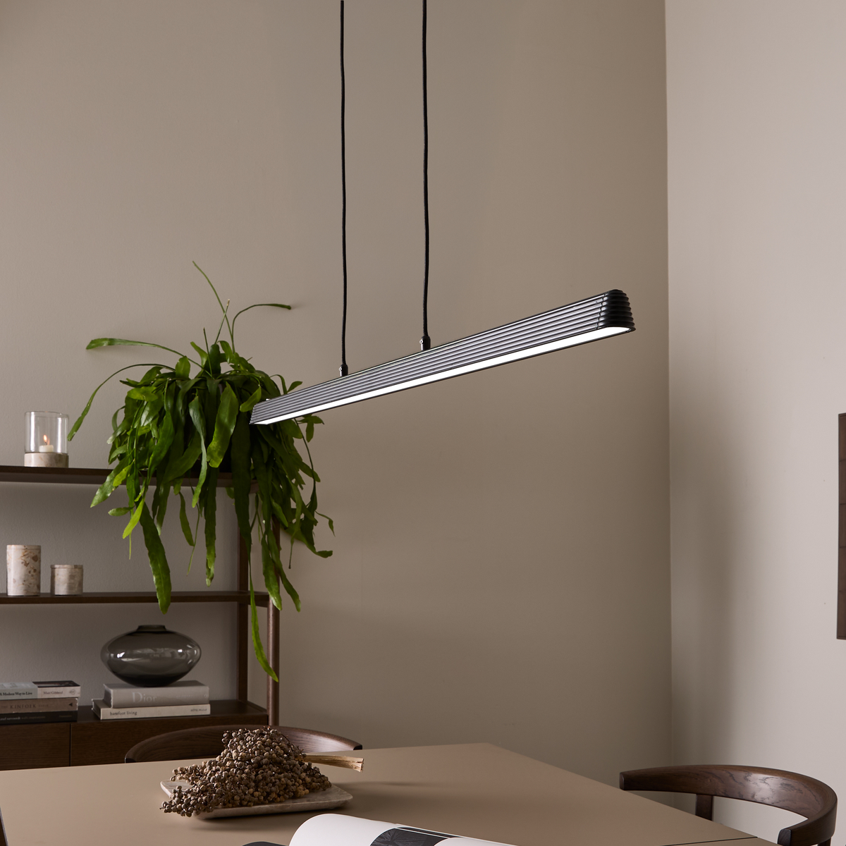 Smal lineær pendellampe i sort metal hænger over et beige spisebord med træstole, plante og reol med dekoration.