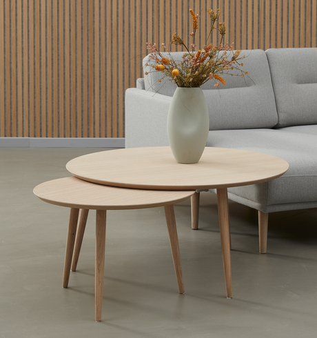 To runde indskudsborde i lyst træ med en vase med tørrede blomster foran en grå sofa.