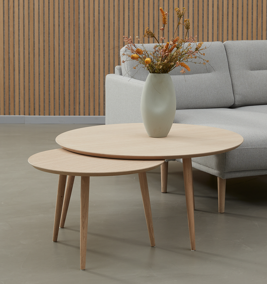 To runde indskudsborde i lyst træ med en vase med tørrede blomster foran en grå sofa.