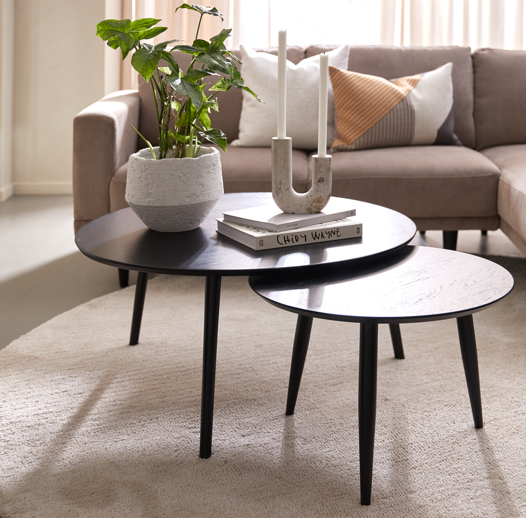 To runde sorte sofaborde med en plante i potte, en U‑formet lysestage med to lys og bøger “Chidy Wayne”, foran en beige sofa med puder i en lys stue.