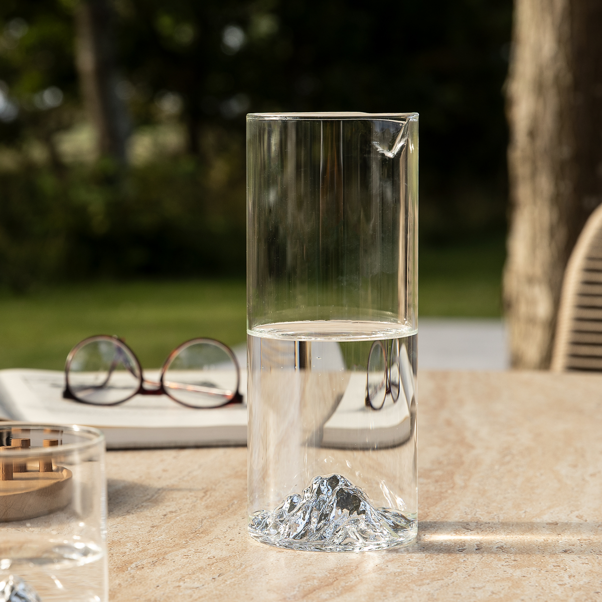 Klar glas-karaff med bjerglignende bund, halvfyldt med vand på et udendørs bord; uskarpe briller og bog i baggrunden.