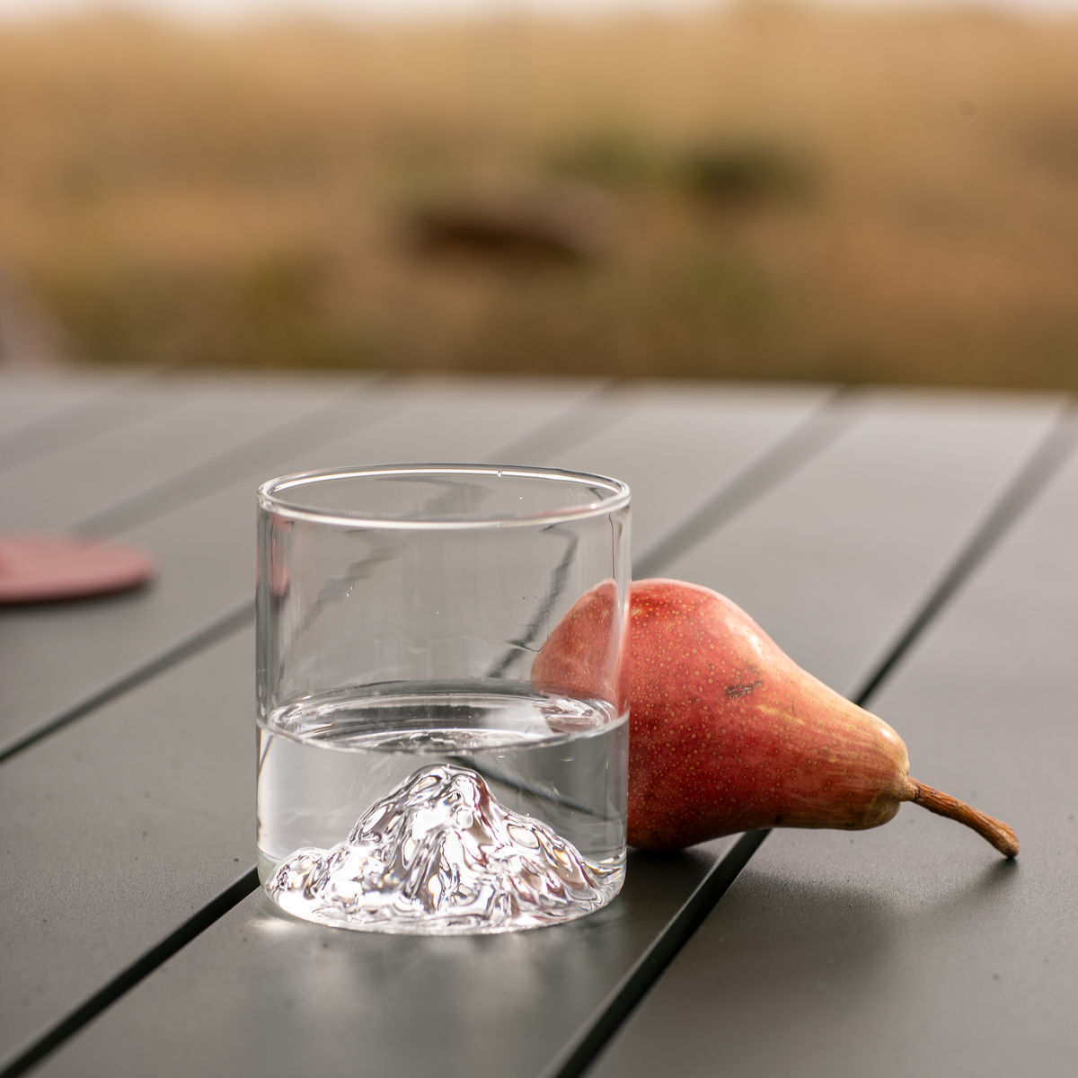 Et glas vand med skulptureret bund ved siden af en rød pære på et udendørs bord.