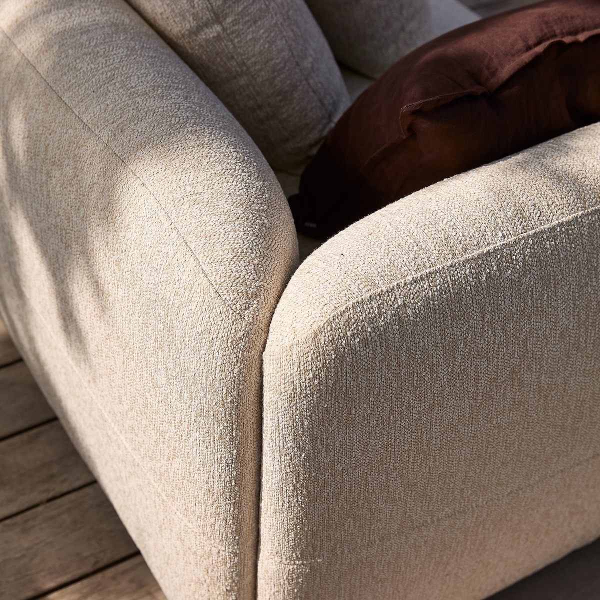 Nærbillede af beige bouclé-sofa med afrundede armlæn og brun pude på træterrasse i dagslys.