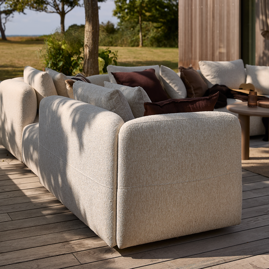 Beige modulsofa udendørs med bløde puder på en træterrasse, rundt sofabord og solpletter, med grøn have i baggrunden.