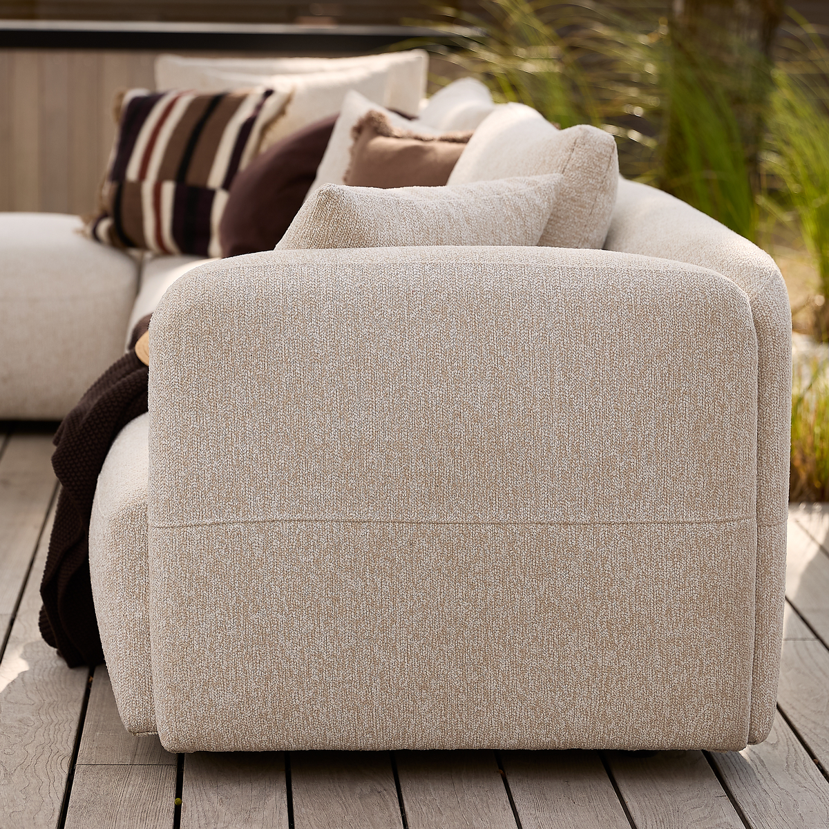 Beige modulsofa på træterrasse med neutrale puder og brun plaid, udendørs i dagslys.