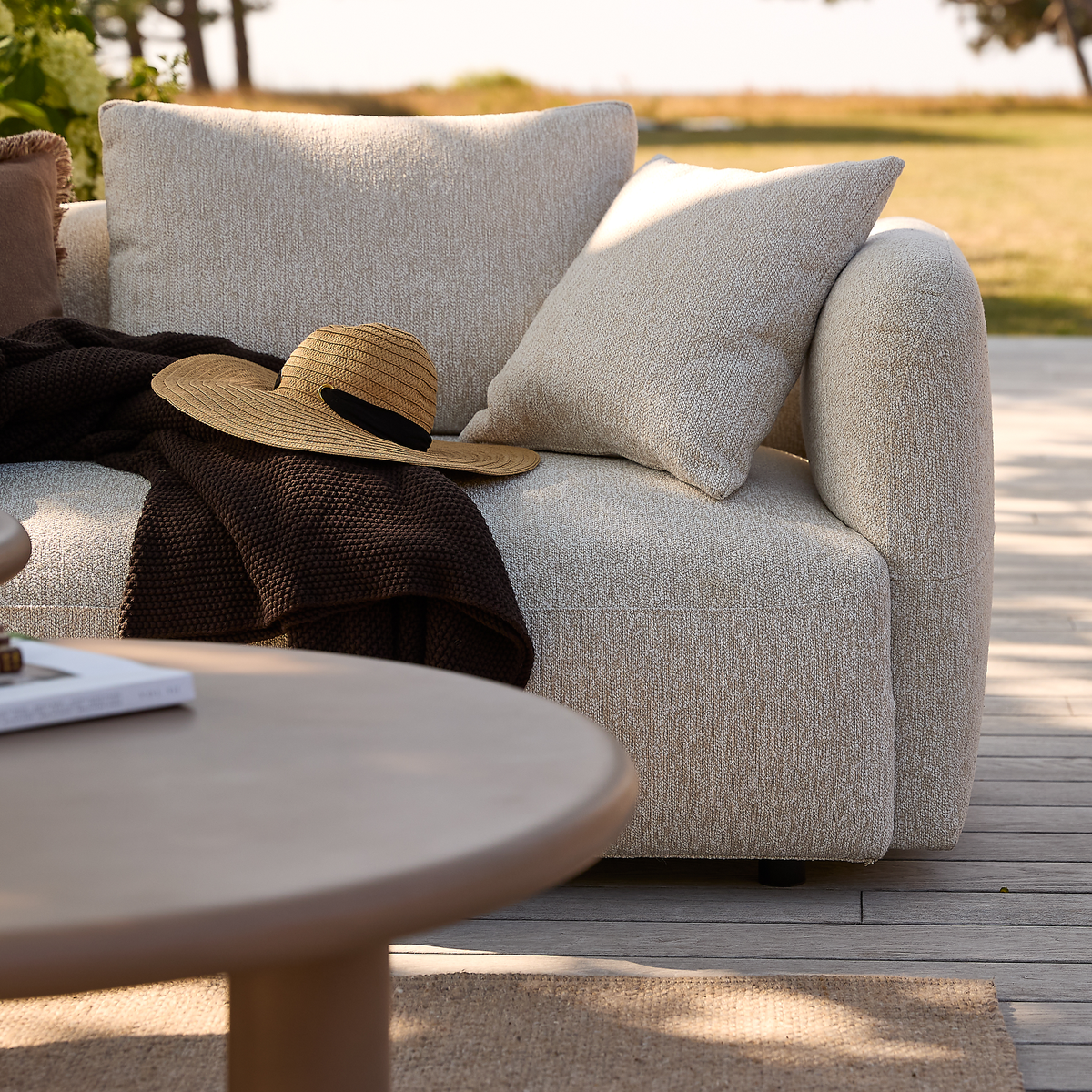 Beige udendørs sofa med puder, brun plaid og stråhat på en træterrasse ved siden af et rundt sofabord i dagslys.