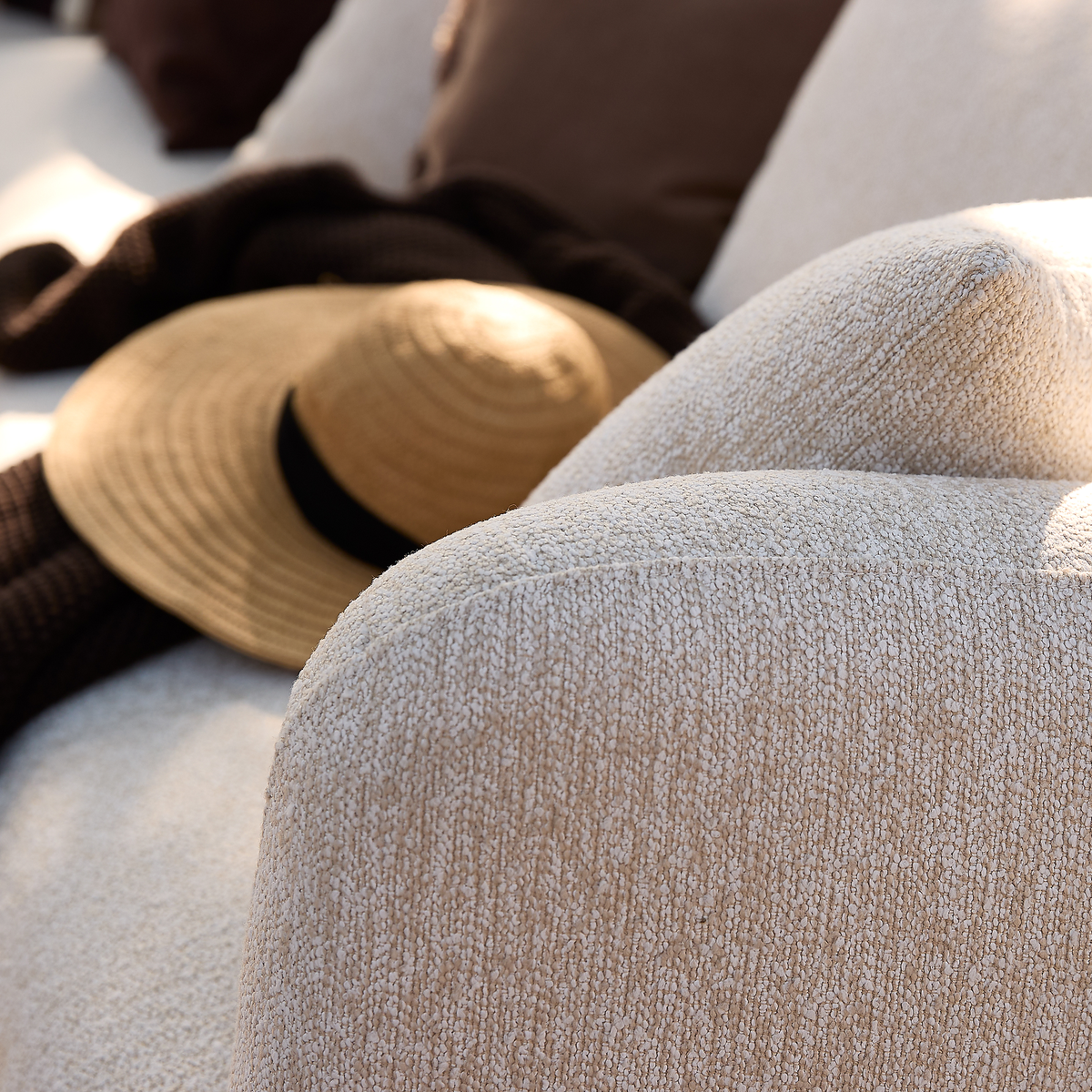 Nærbillede af et beige, tekstureret sofa-armlæn med en stråhat og mørkt strikket plaid på puderne i varmt sollys.