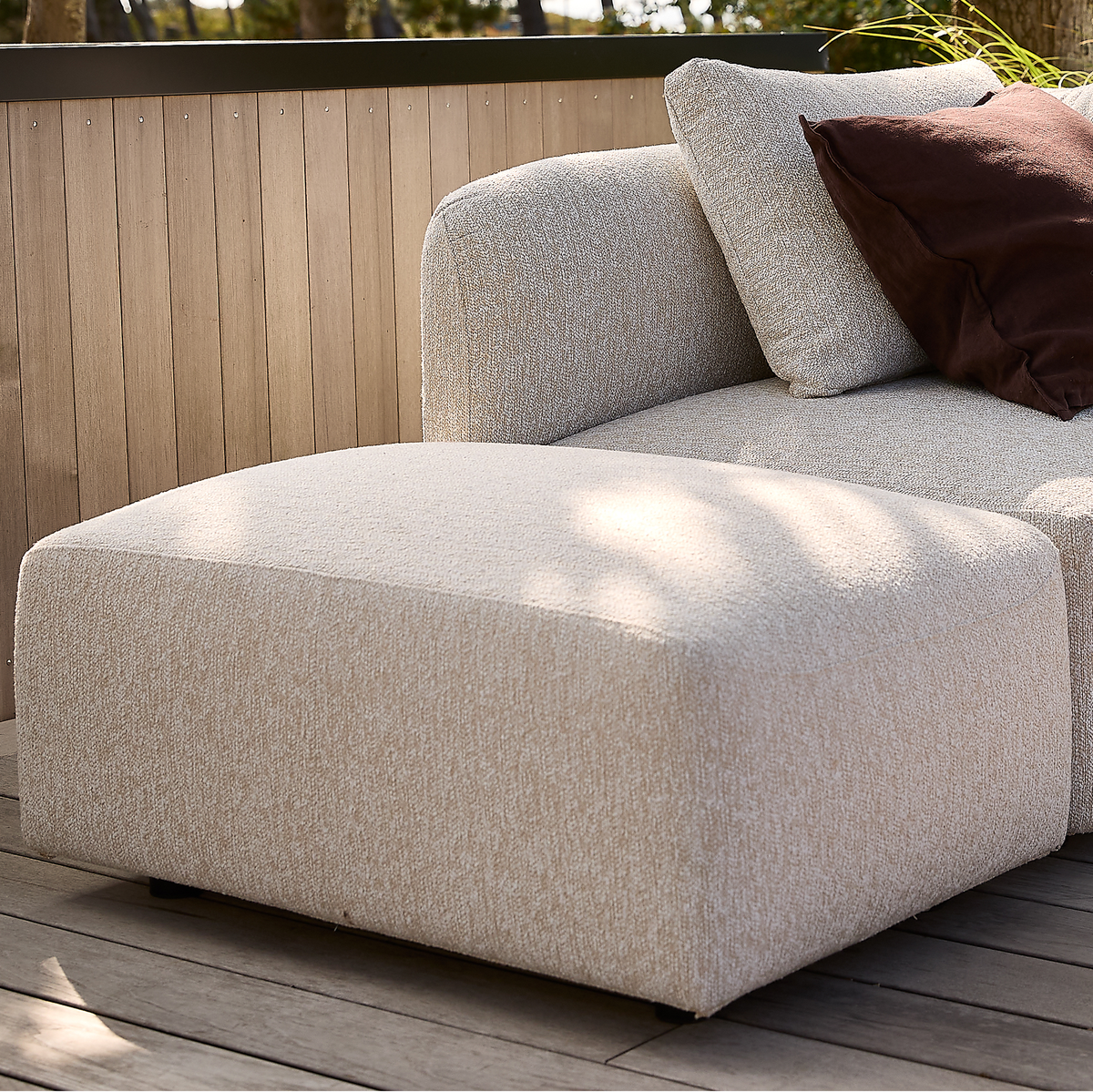 Beige modulsofa med ottoman på en træterrasse, med brun pyntepude.