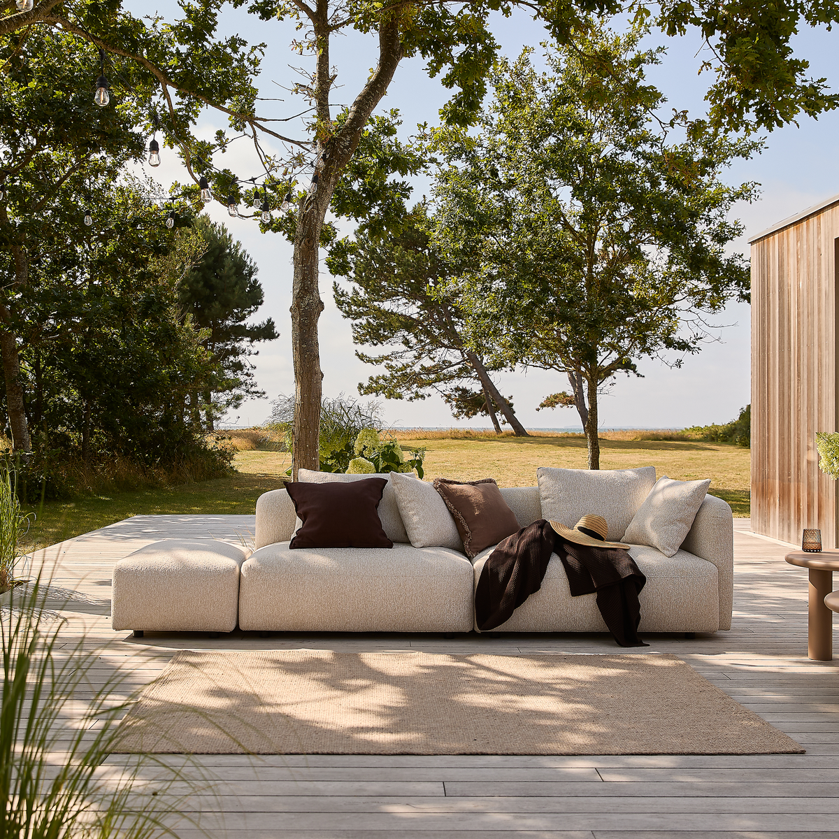 Beige modulsofa udendørs med brune og hvide puder på en træterrasse, lys måtte og lille sidebord; træer og eng i baggrunden i dagslys.