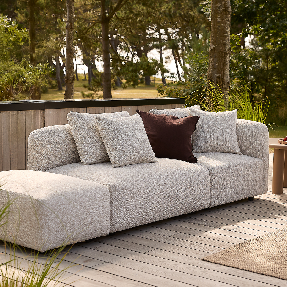 Lys beige modulsofa med chaiselong på træterrasse, fire puder hvoraf én mørkebrun accentpude, omgivet af grønne træer i dagslys.