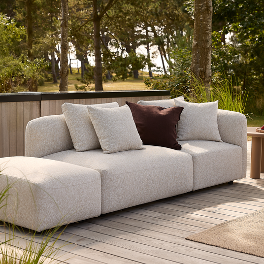 Lys beige modulsofa med chaiselong på træterrasse, fire puder hvoraf én mørkebrun accentpude, omgivet af grønne træer i dagslys.
