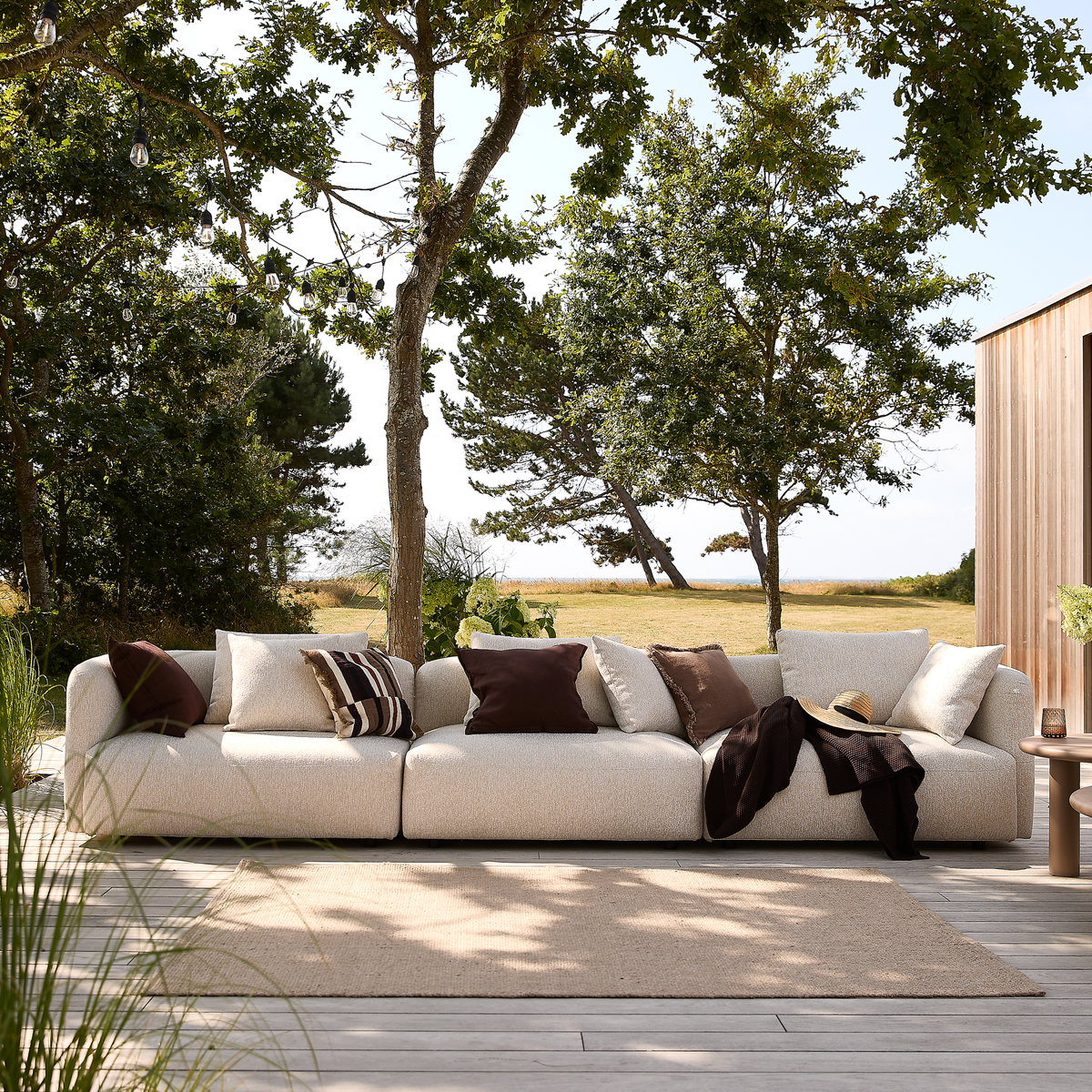 Beige modulsofa på træterrasse udendørs med brune og lyse puder, tæppe og sidebord i træernes skygge med udsigt mod eng og vand.