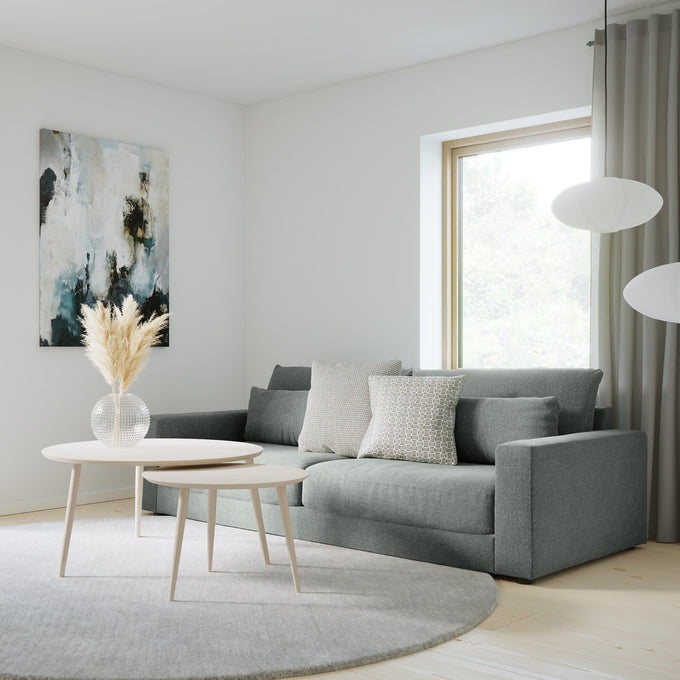 Lyst skandinavisk stue med grå sofa, to runde sofaborde, lysgråt rundt tæppe, abstrakt vægkunst og to hvide pendellamper; store vinduer med gardiner og en vase med tørrede strå på bordet.