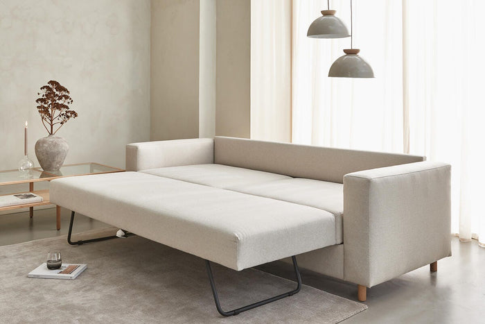 Beige sovesofa trukket ud i minimalistisk stue med to pendellamper, lette gardiner og glasbord med vase og stearinlys.