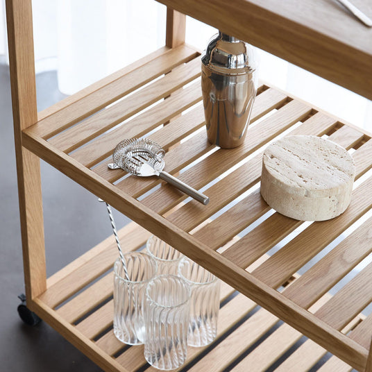 Nærbillede af en barvogn i lyst træ med en rustfri cocktailshaker, si/strainer, rund sten-underlag og riflede glas, i blødt dagslys ved en hvid gardin.
