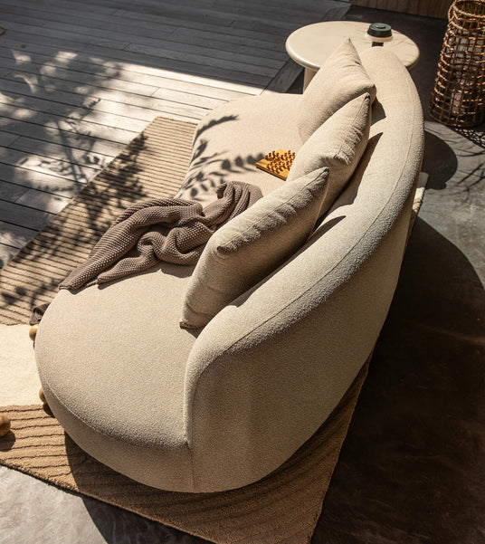 Beige, rund sofa udendørs på træterrasse med puder, brun strikket plaid, rundt sidebord og kurv i varmt dagslys.