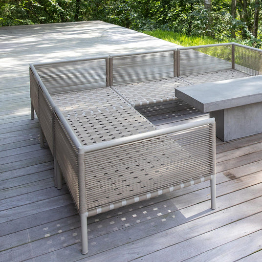 Udendørs terrasse med moderne loungemøbler i snor og et betonbord på trædæk, omgivet af grøn skov.
