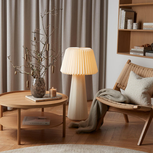 Hyggekrog i stuen i skandinavisk stil med fletlounge-stol, pude og plaid, plisseret gulvlampe og rundt sofabord med vase med grene og et tændt duftlys; bøgerne KINFOLK HOME og New Nordic Houses ses på hylden.