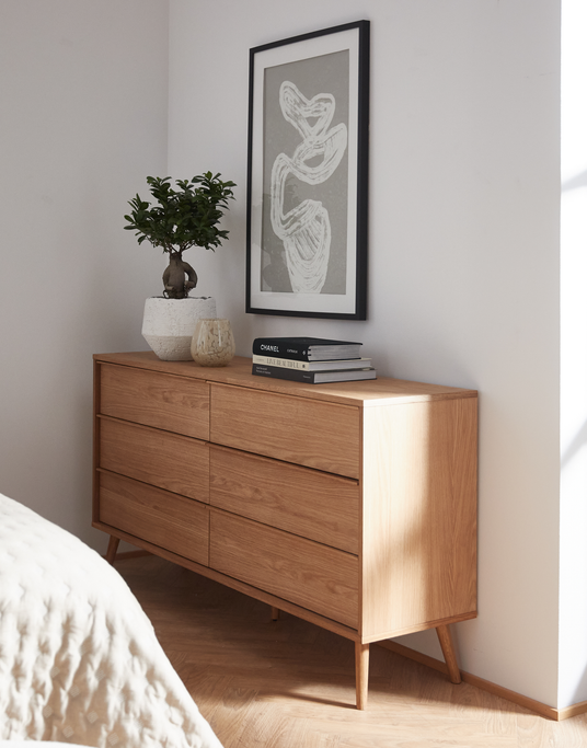 Kommode i lys eg med seks skuffer i et lyst soveværelse; ovenpå står en bonsai i hvid potte, et lys og en bogstak med blandt andet Chanel, under en abstrakt indrammet plakat.