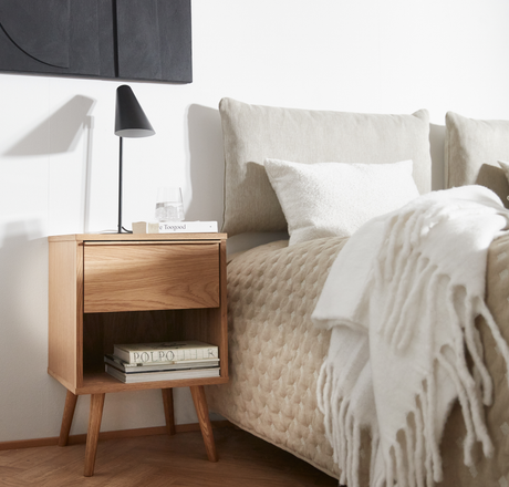 Soveværelse med natbord i træ, sort bordlampe, glas vand og bøgerne ”Faye Toogood” og ”POLPO”; beige sengetæppe med hvid frynset plaid.