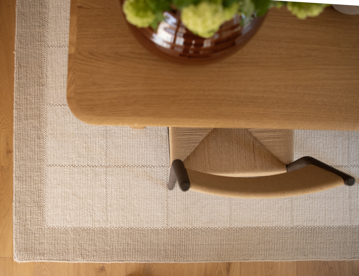 Set ovenfra: et træbord med vase med grønne blomster, en stol med fletsæde og et lyst beige tæppe med kant på trægulv.