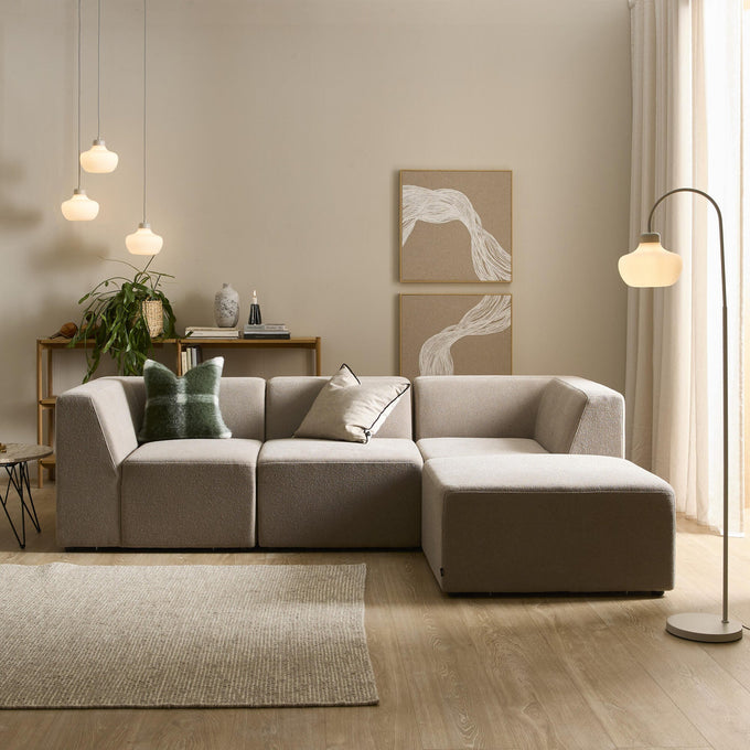 Lyst stue i neutrale toner med beige modulsofa med chaiselong, to puder, gulvlampe og tre pendler, vægkunst, tæppe og et konsolbord med plante og bøger.