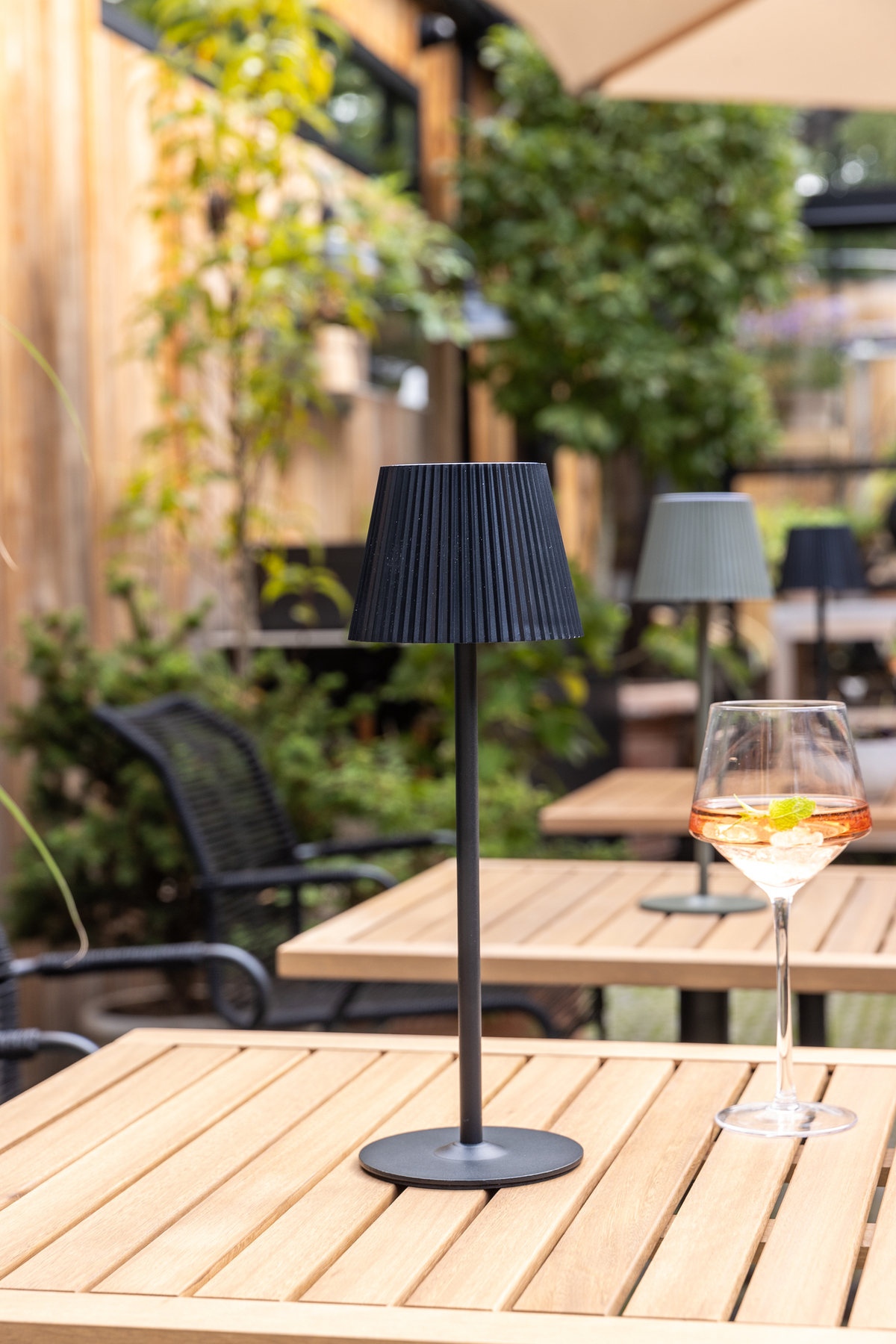 Sort bærbar bordlampe på træbord på terrasse ved siden af et vinglas med is og mynte.